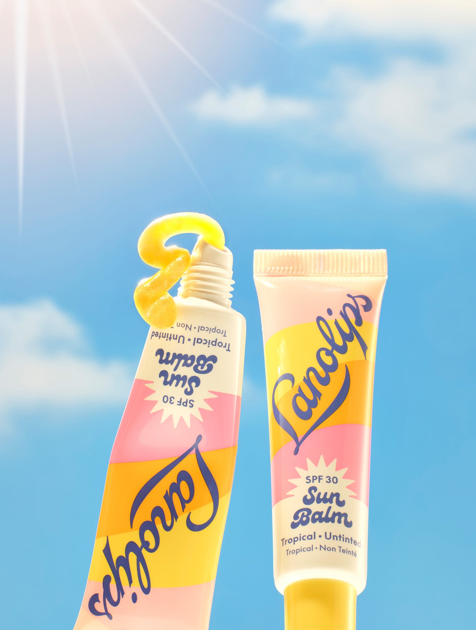 Set against a radiant blue sky with a shining sun, two tubes of Lanolips Sun Balm SPF 30 Tropical lip balm are featured - one standing upright and the other squeezed to reveal a vibrant, glossy balm shaped like a “3” to highlight its SPF 30 protection. This creamy, radiant formula is packed with hydrating lanolin, SPF protection, and a luminous sheen.