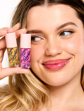 Smiling model holding two Lanolips Hyaluronic Lip Oil shades, highlighting the product's sleek pink packaging. The lip oils, infused with lanolin, hyaluronic acid, and vitamin E, deliver a glossy and nourishing finish for soft, hydrated lips