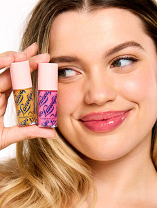 Smiling model holding two Lanolips Hyaluronic Lip Oil shades, highlighting the product's sleek pink packaging. The lip oils, infused with lanolin, hyaluronic acid, and vitamin E, deliver a glossy and nourishing finish for soft, hydrated lips