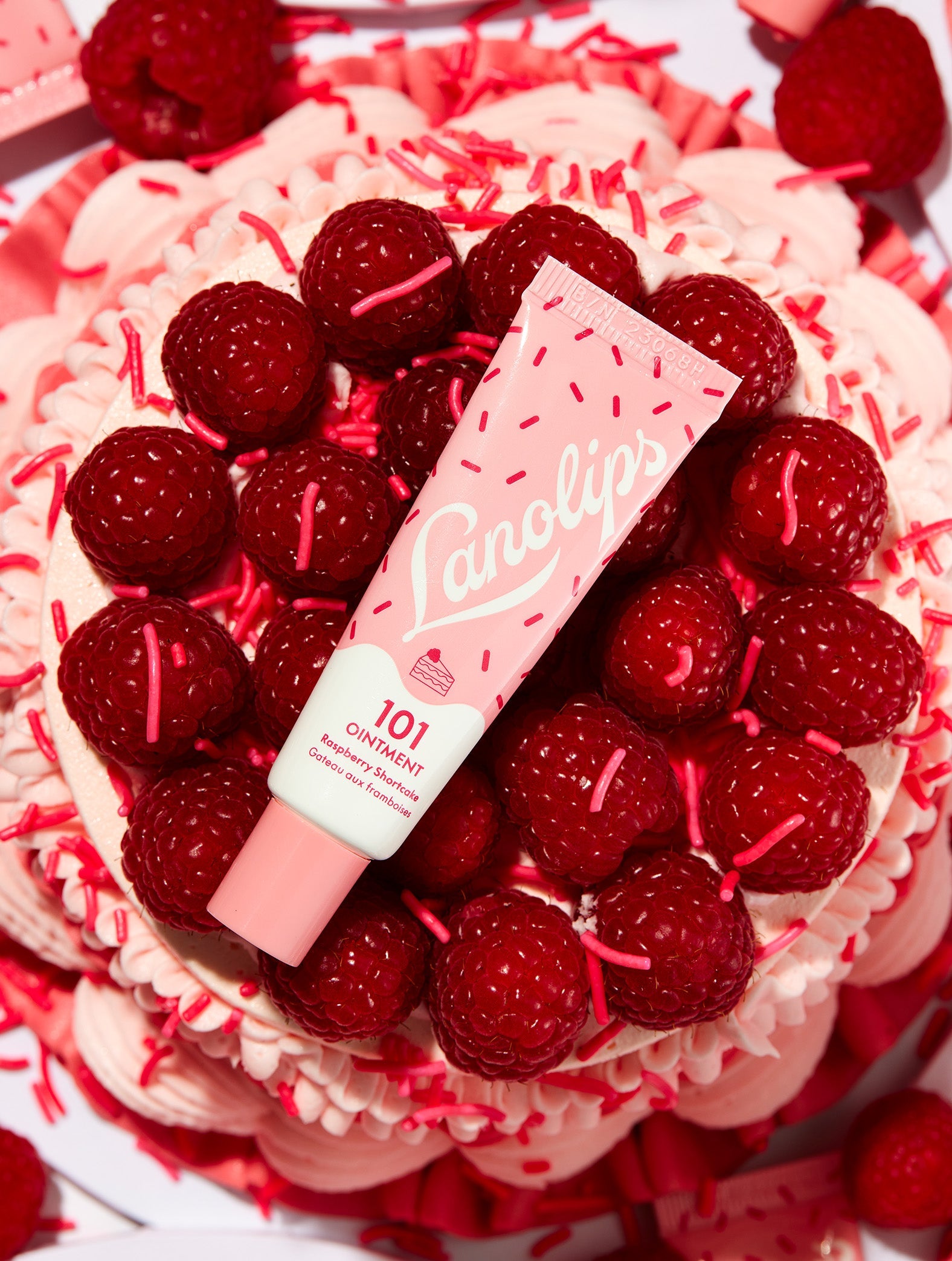 Flat lay of Lanolips 101 Ointment Multi-Balm in Raspberry Shortcake, placed on a swirl of pink frosting, fresh raspberries and sprinkles. This all-natural balm blends juicy raspberries with buttery shortcake for sweet hydration and a nourishing moisture boost.