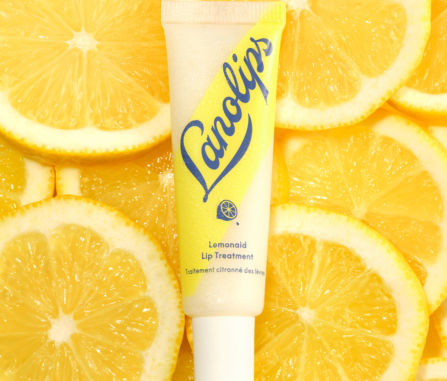 A tube of Lanolips Lemonaid Lip Treatment sits among fresh, bright yellow lemon slices. The packaging features a yellow and white gradient with bold blue Lanolips lettering and a small lemon icon. The lip treatment, infused with rich lanolin, all-natural lemon oil, vitamin e and a hint of shimmer, deeply nourishes, gently exfoliates, and leaves lips feeling soft, smooth, and hydrated with a glossy finish.