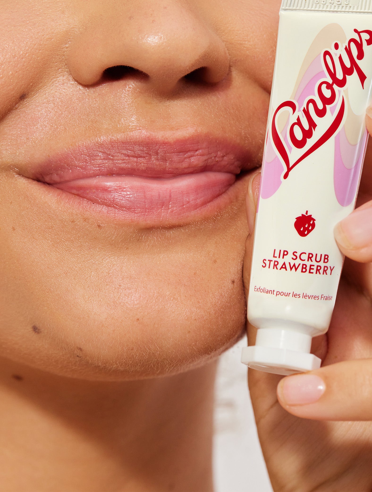 Model's hand holding a lanolips lip scrub in strawberry against her face.