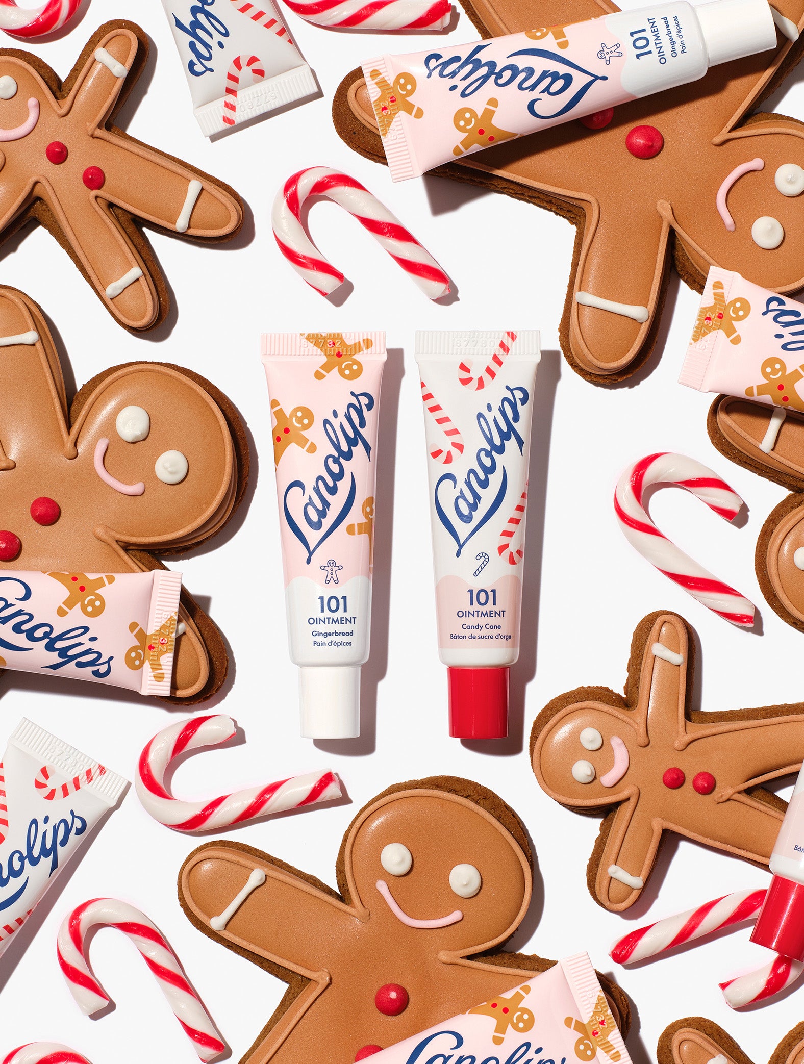 Lanolips 101 Ointment Multi-Balm Gingerbread & Candy Cane centred among gingerbread cookies, candy canes and balm tubes, from the Lanolips Holiday Gingerbread House Lip Duo. Hydrating lanolin, vitamin E and natural flavours keep lips soft and festive.