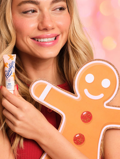 Model holding a cut out of the gingerbread man cut out and a Lanolips 101 Ointment Multi-Balm Gingerbread against a colourful background.